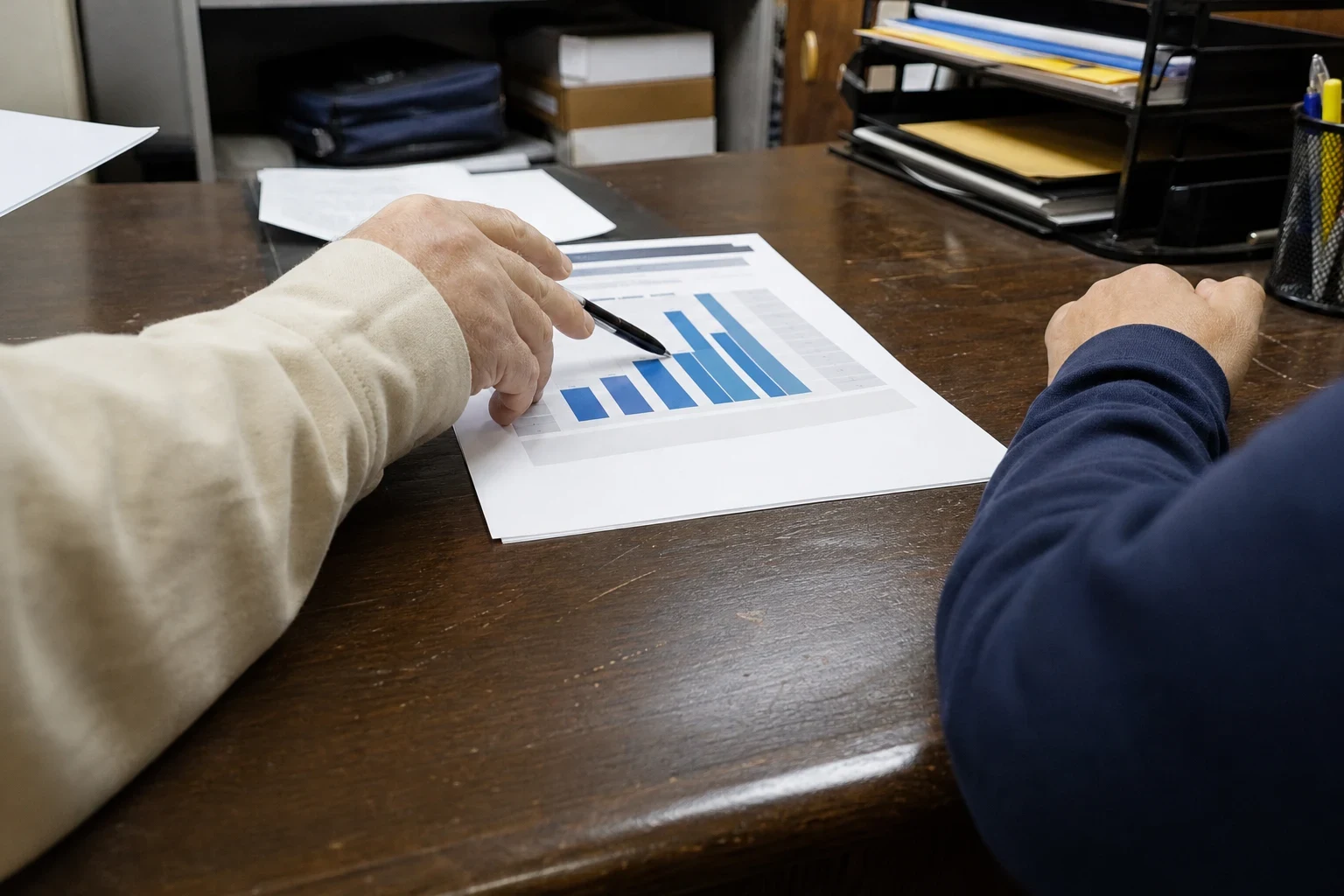Hand holding a pen points to a blue bar chart on a printed sheet across a worn wooden desk, with a second person's arm in a navy sleeve and file trays nearby